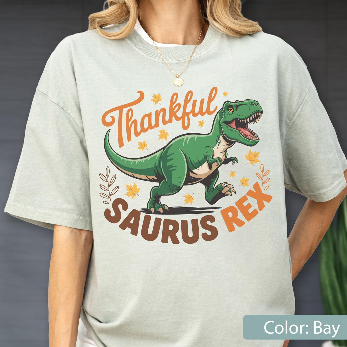 May include: A light green t-shirt featuring a green Tyrannosaurus Rex illustration with the text "Thankful Saurus Rex" in orange and brown. The design includes fall leaves. The shirt is a casual style.