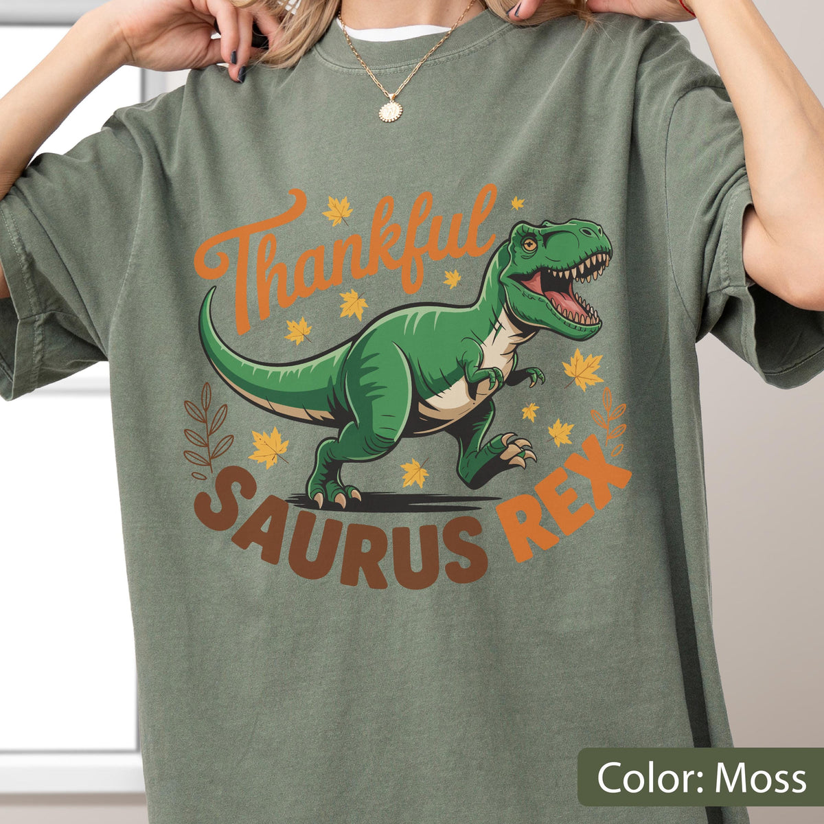 May include: Moss green t-shirt featuring a green Tyrannosaurus Rex dinosaur with the words "Thankful Saurus Rex" in orange and brown. The design includes fall leaves and a leafy border. The shirt is a solid color.