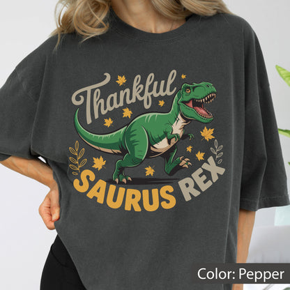 May include: Pepper-colored t-shirt featuring a green Tyrannosaurus Rex illustration with the text "Thankful Saurus Rex." The design includes fall leaves and a decorative border, perfect for the autumn season.