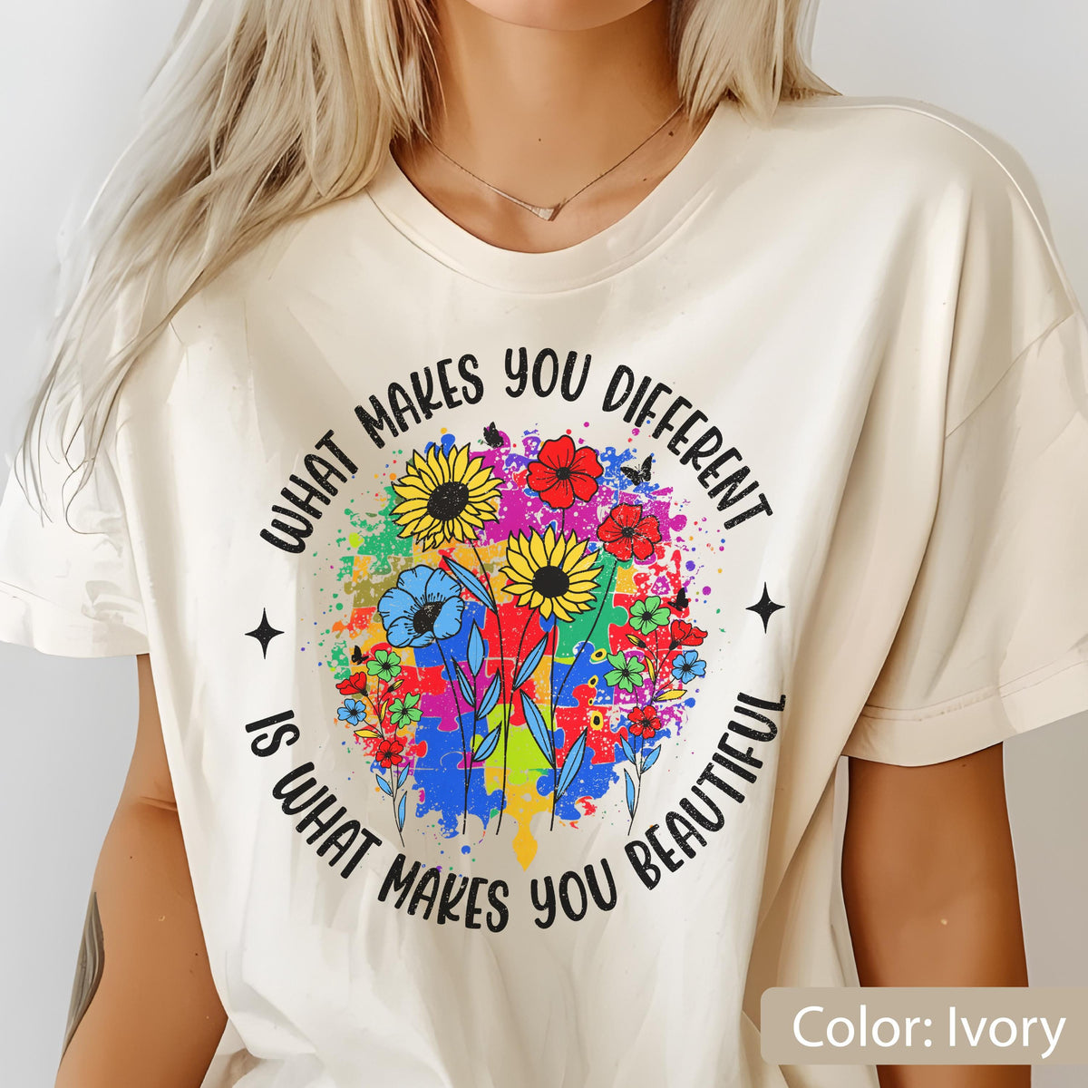 May include: Ivory-colored t-shirt with a colorful floral design. The design includes sunflowers, red flowers, and blue flowers, with a puzzle piece background. The text reads: "What makes you different is what makes you beautiful."