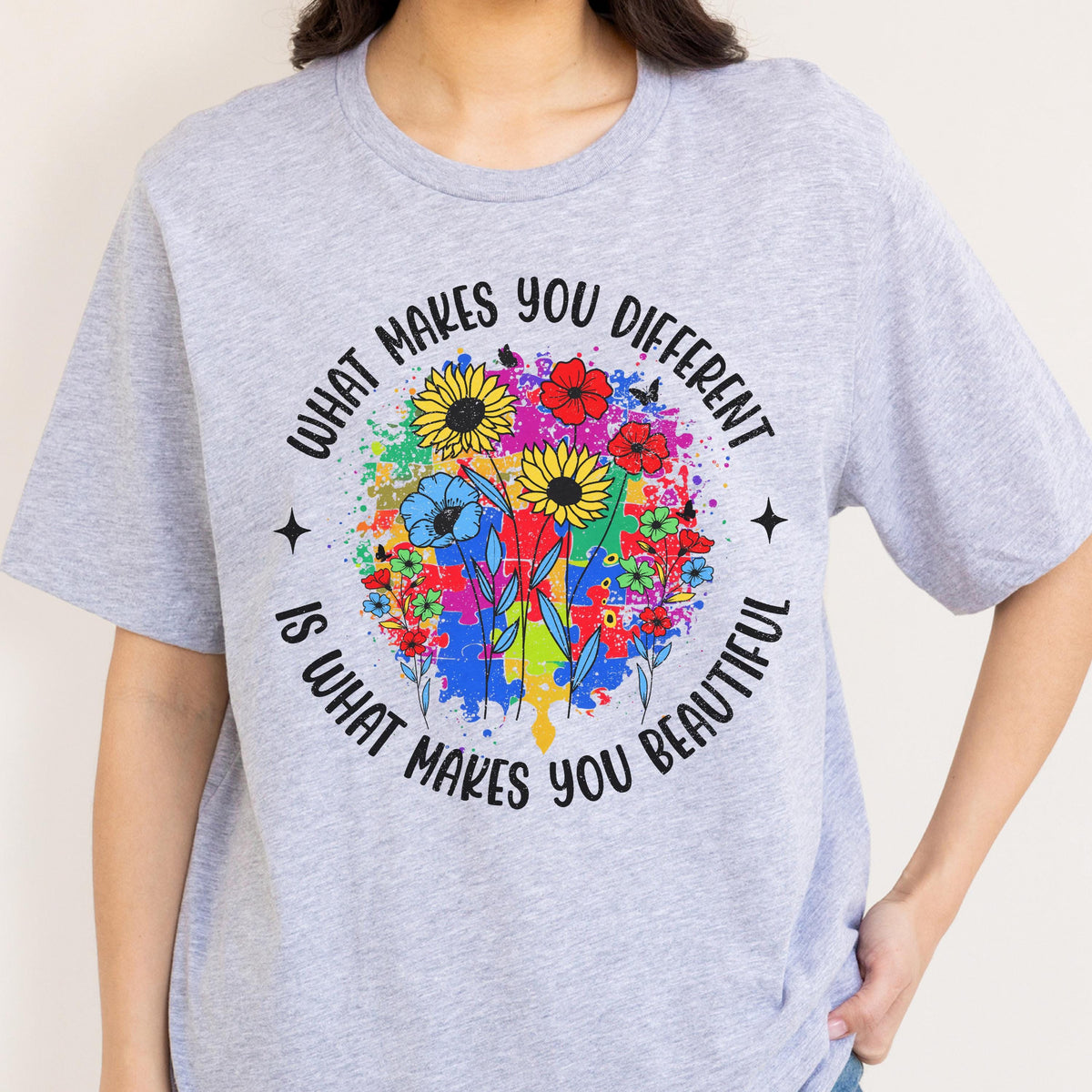May include: A light gray t-shirt featuring a colorful floral design with the text "WHAT MAKES YOU DIFFERENT IS WHAT MAKES YOU BEAUTIFUL" in a circular pattern. The design includes sunflowers, red flowers, and blue flowers.