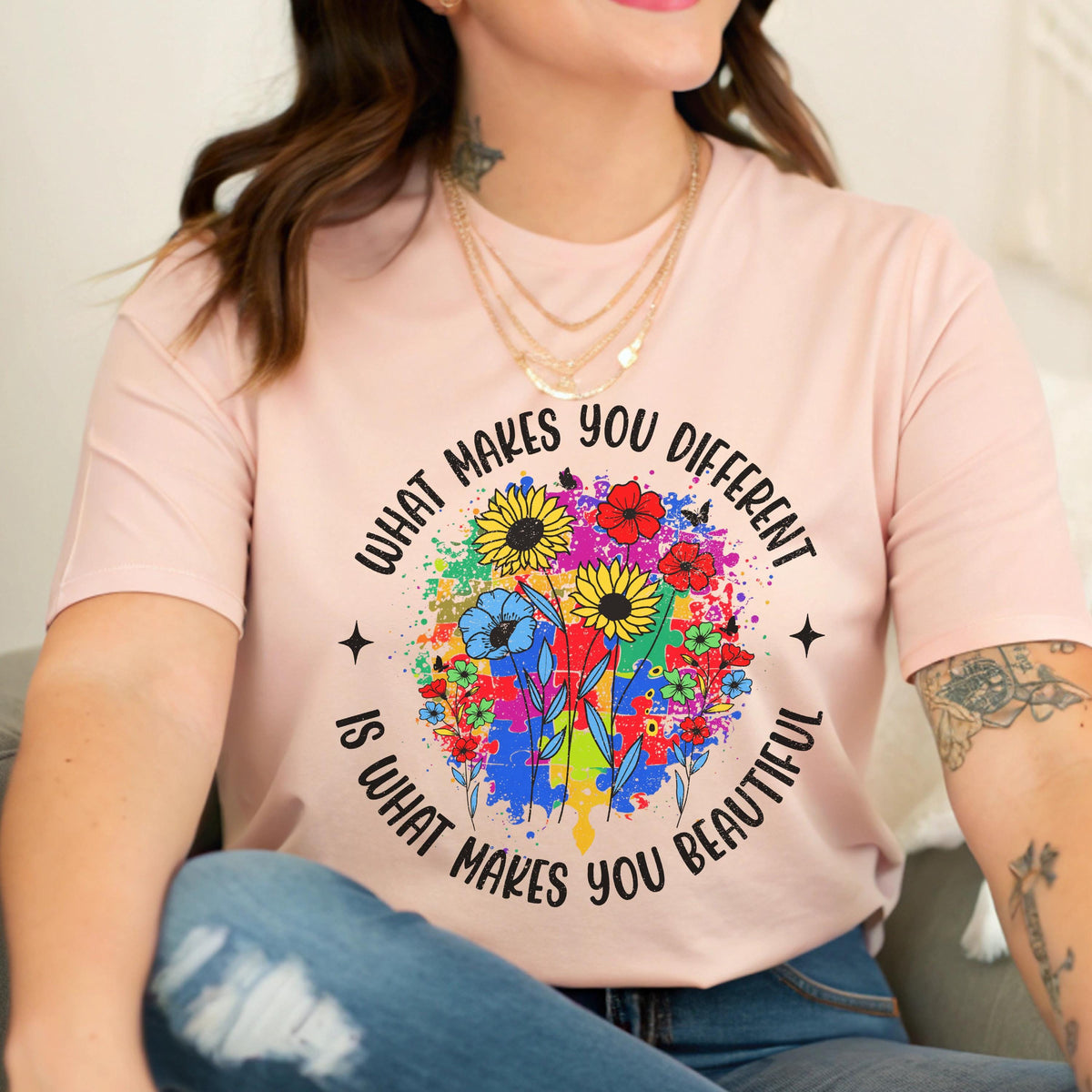 May include: A light pink t-shirt with the text "WHAT MAKES YOU DIFFERENT IS WHAT MAKES YOU BEAUTIFUL" encircling a colorful floral design. The design includes sunflowers, blue flowers, and puzzle pieces.