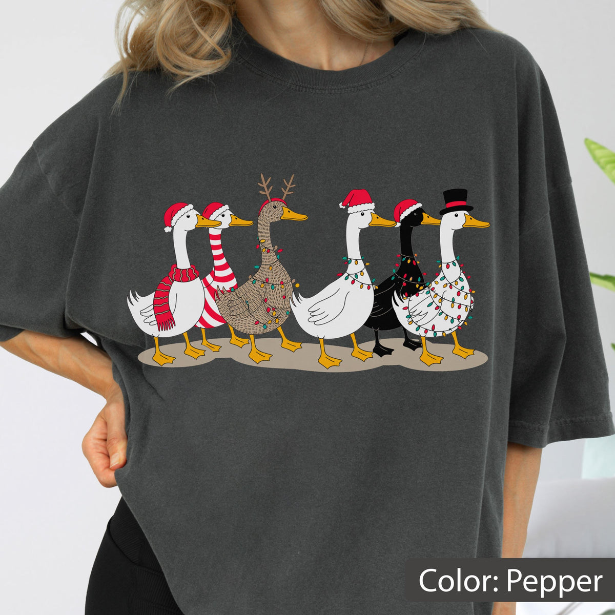 May include: A charcoal gray t-shirt featuring a graphic of six geese walking in a line. The geese are decorated with Christmas elements including Santa hats, a top hat, reindeer antlers, striped scarves, and colorful string lights wrapped around them.