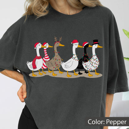 May include: A charcoal gray t-shirt featuring a graphic of six geese walking in a line. The geese are decorated with Christmas elements including Santa hats, a top hat, reindeer antlers, striped scarves, and colorful string lights wrapped around them.