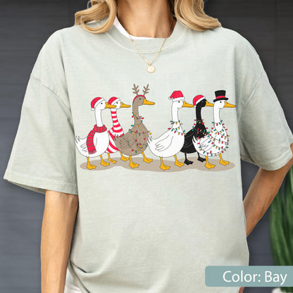 May include: A light sage green t-shirt featuring a graphic print of six ducks in a row. The ducks are decorated with various holiday accessories including Santa hats, a top hat, a red and white striped scarf, reindeer antlers, and colorful string lights.
