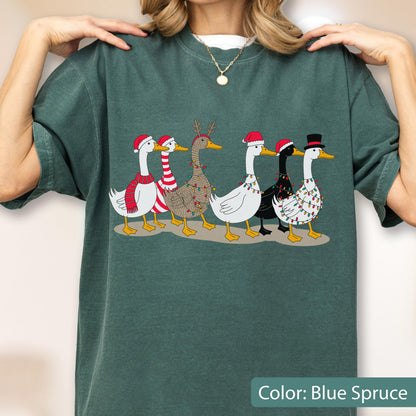 May include: A dark green t-shirt featuring a graphic of six geese walking in a line. The geese are decorated with Christmas elements including Santa hats, a top hat, a red and white striped scarf, reindeer antlers, and colorful string lights.