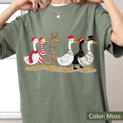 May include: A moss green t-shirt featuring a graphic print of six ducks in a row. The ducks are decorated for the holidays with Santa hats, a top hat, red and white striped scarves, and colorful string lights wrapped around their bodies.