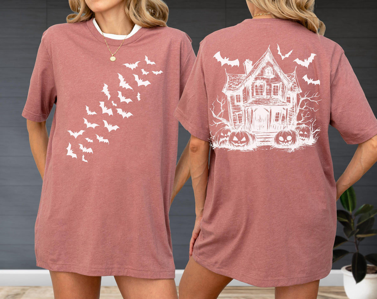 May include: A dusty rose-colored t-shirt with a white bat design on the front and a haunted house graphic on the back. The front features a diagonal pattern of bats. The back has a spooky house, bats, and jack-o'-lanterns.