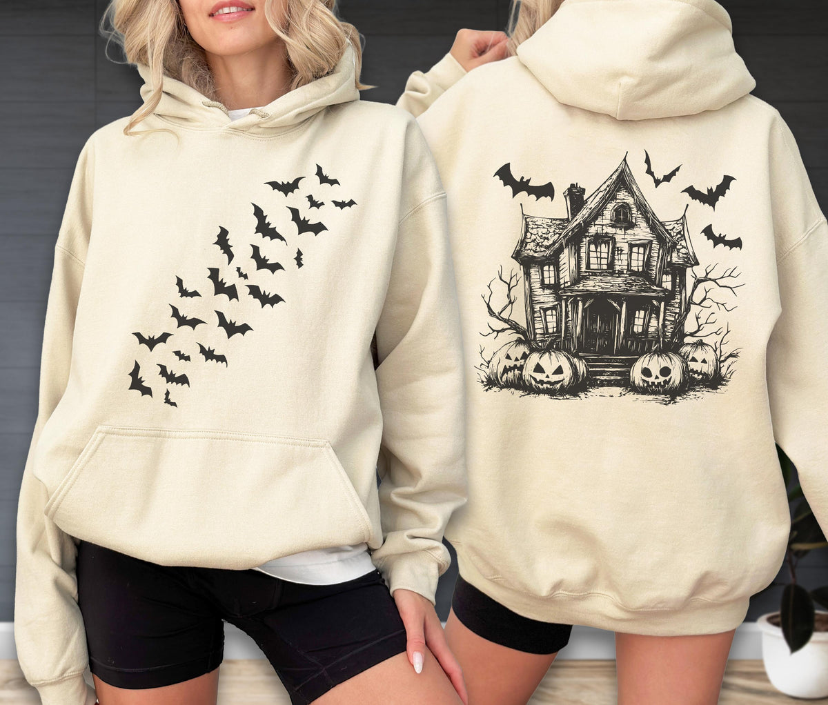 May include: A cream-colored hooded sweatshirt featuring a black graphic of a swarm of bats on the front chest. The back of the hoodie displays a detailed black illustration of a haunted house surrounded by jack-o'-lanterns and flying bats.