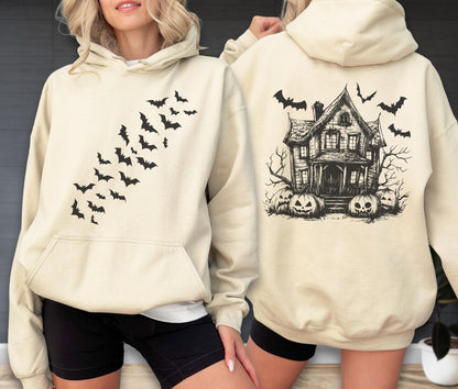 May include: A cream-colored hooded sweatshirt featuring a black graphic of a swarm of bats on the front chest. The back of the hoodie displays a detailed black illustration of a haunted house surrounded by jack-o'-lanterns and flying bats.