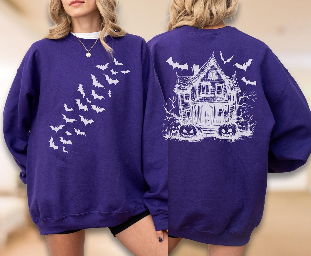 May include: A deep purple crewneck sweatshirt featuring a white graphic print. The front displays a diagonal pattern of flying bats. The back showcases a detailed illustration of a haunted house surrounded by bare trees and carved jack-o'-lanterns.