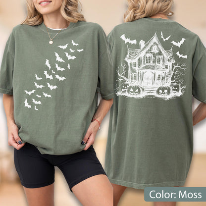 May include: An olive green t-shirt featuring a white graphic of a swarm of bats on the front. The back displays a white line-art illustration of a haunted house surrounded by bare trees, jack-o'-lanterns, and flying bats against the fabric.