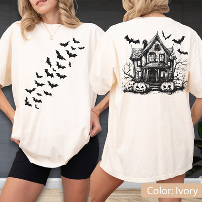 May include: An ivory t-shirt featuring a black graphic of a swarm of bats on the front chest area. The back of the t-shirt displays a detailed black ink illustration of a haunted house surrounded by bare trees and several carved jack-o'-lanterns.