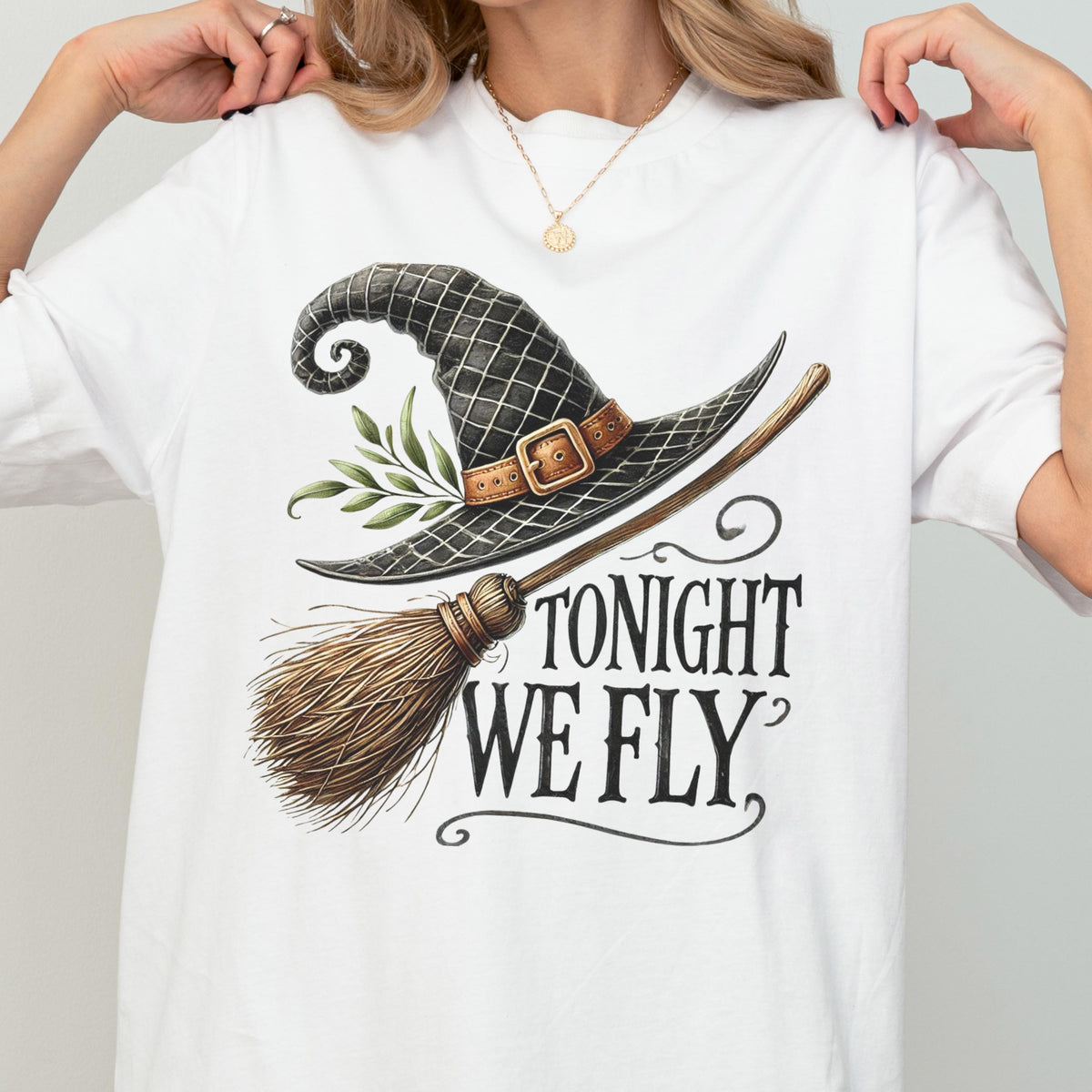 May include: A white t-shirt featuring a graphic print of a black witch hat with a brown buckle and green leaves, paired with a wooden broomstick. The text "TONIGHT WE FLY" is printed in a black, stylized serif font below the hat and broom.