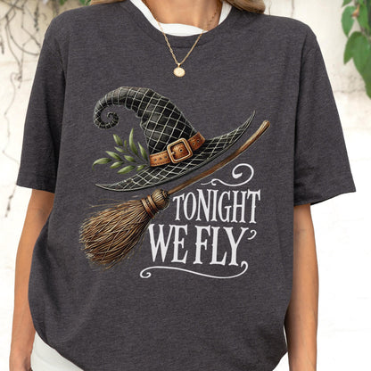 May include: A charcoal gray t-shirt featuring a graphic of a black witch hat with a brown buckle and green leaves, paired with a wooden broomstick. The text TONIGHT WE FLY is printed in white lettering below the hat and broomstick illustration.