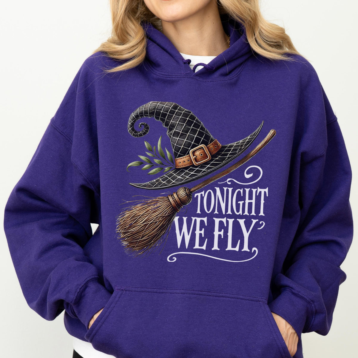 May include: A deep purple hooded sweatshirt featuring a graphic print of a black witch hat with a brown buckle and green leaves, paired with a wooden broomstick. The text TONIGHT WE FLY is printed in white lettering below the graphic design.