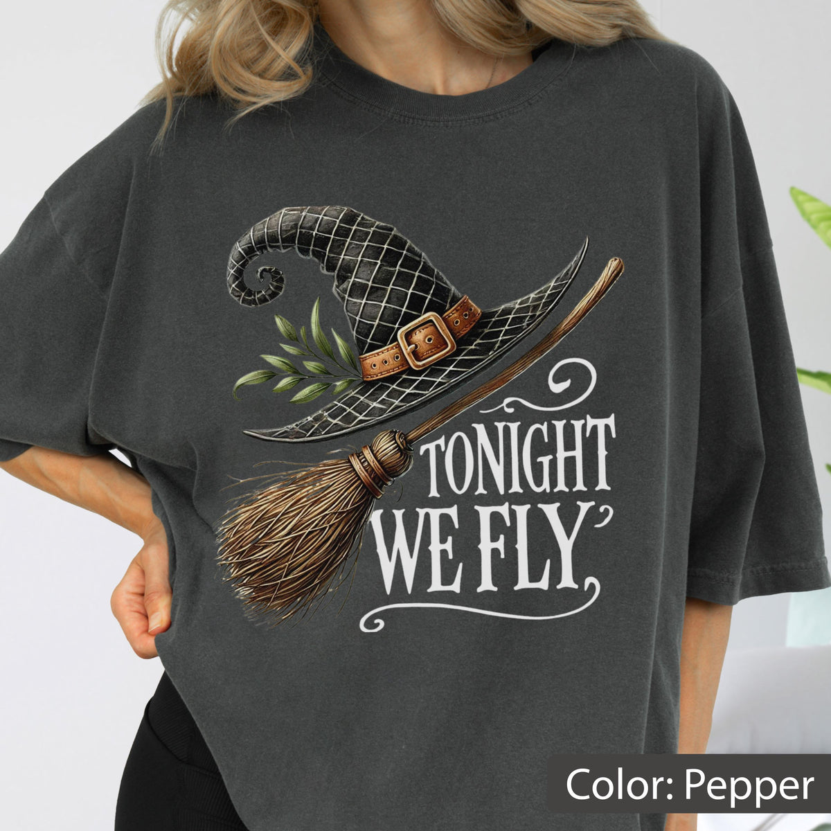 May include: A charcoal gray t-shirt featuring a graphic print of a black witch hat with a brown buckle and green leaves, paired with a wooden broomstick. The text TONIGHT WE FLY is printed in white in a decorative serif font below the graphic.