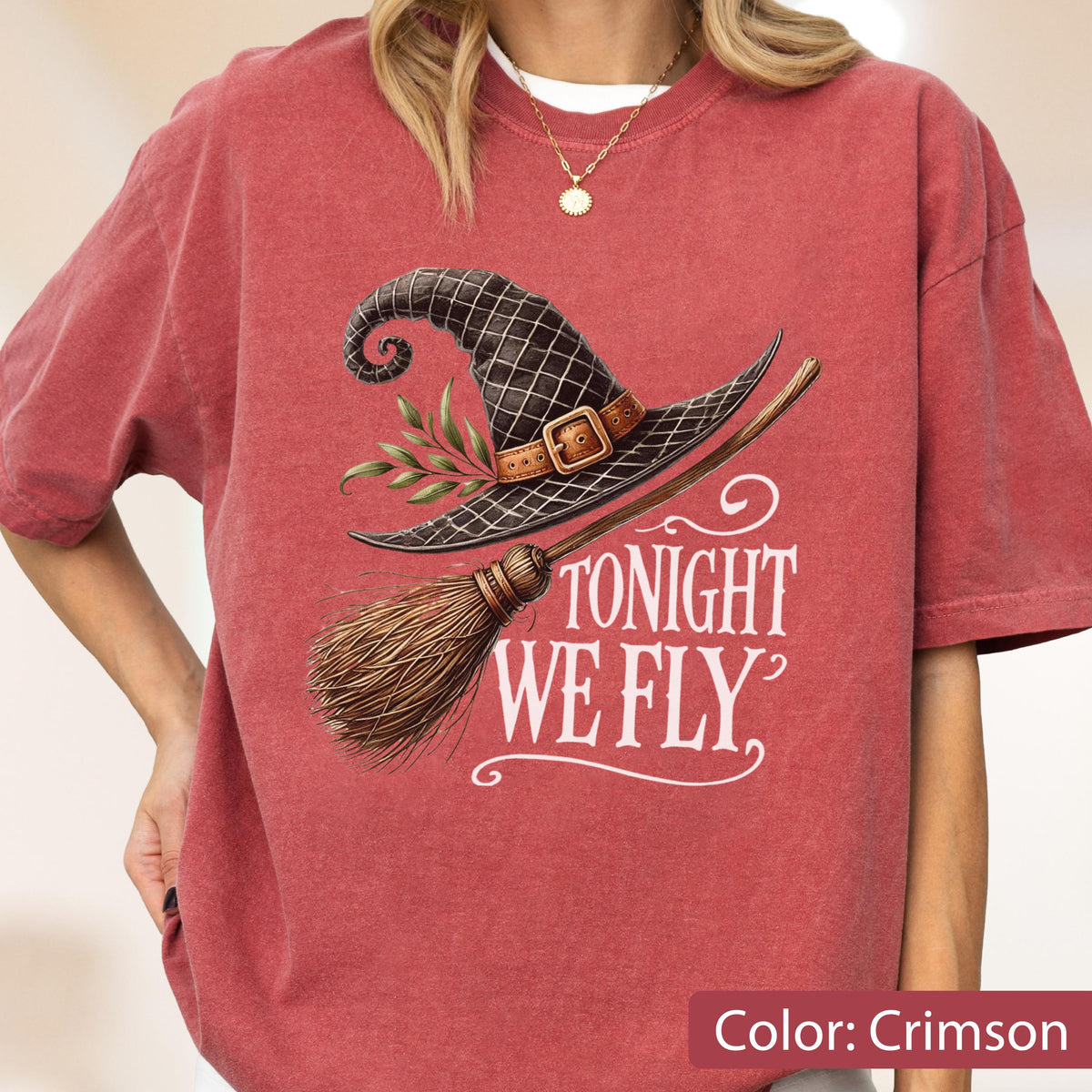 May include: A crimson short-sleeve t-shirt featuring a graphic of a black witch hat with a brown buckle and green leaves, paired with a wooden broomstick. The text TONIGHT WE FLY is printed in white below the hat and broomstick illustration.