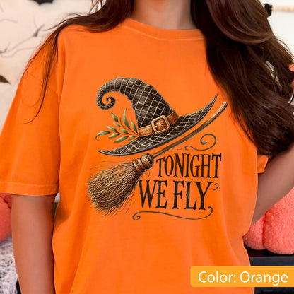 May include: An orange t-shirt featuring a graphic print of a dark brown witch hat with a buckle and green leaves, paired with a wooden broomstick. The text "TONIGHT WE FLY" is printed in a black, stylized serif font below the illustration.