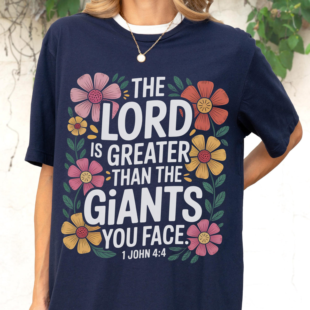May include: A navy blue short-sleeved t-shirt featuring the white text THE LORD IS GREATER THAN THE GIANTS YOU FACE 1 JOHN 4:4. The text is surrounded by a colorful floral border with pink, orange, and yellow flowers and green leaves.