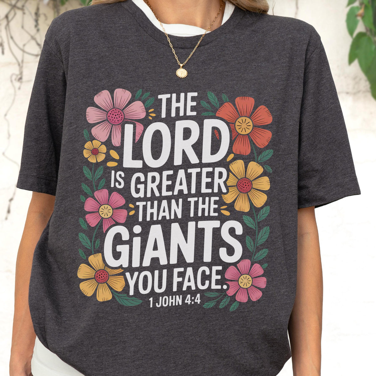 May include: A dark charcoal gray t-shirt featuring a floral border surrounding the white text "THE LORD IS GREATER THAN THE GIANTS YOU FACE. 1 JOHN 4:4". The design includes pink, orange, and yellow flowers with green leaves and stems.
