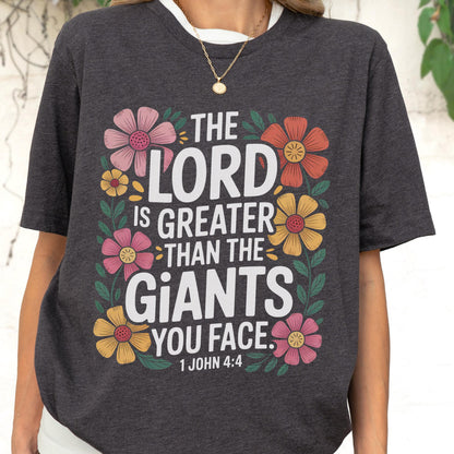May include: A dark charcoal gray t-shirt featuring a floral border surrounding the white text "THE LORD IS GREATER THAN THE GIANTS YOU FACE. 1 JOHN 4:4". The design includes pink, orange, and yellow flowers with green leaves and stems.