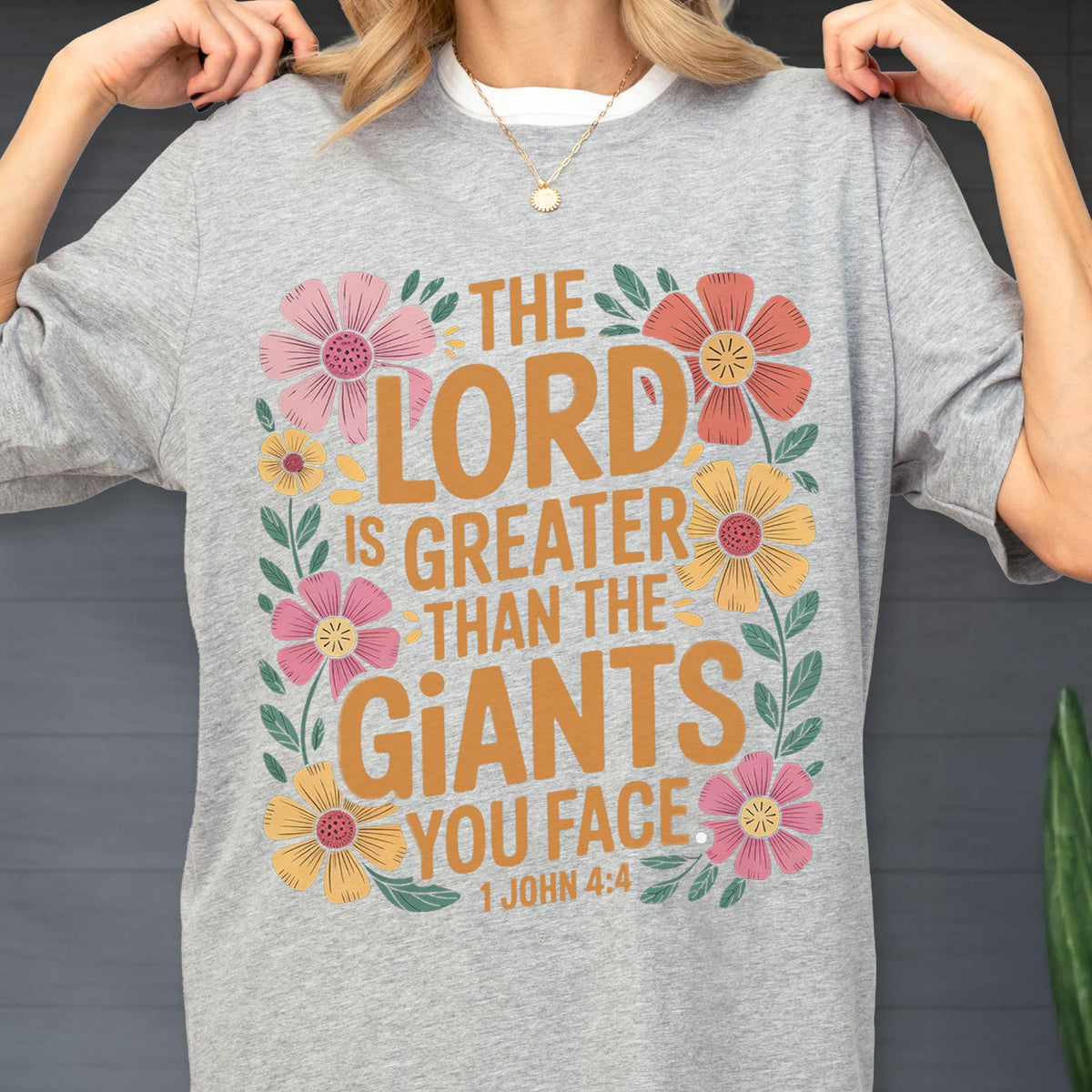 May include: A heather gray t-shirt featuring the text THE LORD IS GREATER THAN THE GIANTS YOU FACE 1 JOHN 4:4 in mustard yellow. The design is surrounded by a floral border with pink, orange, and yellow flowers and green leaves on the front.