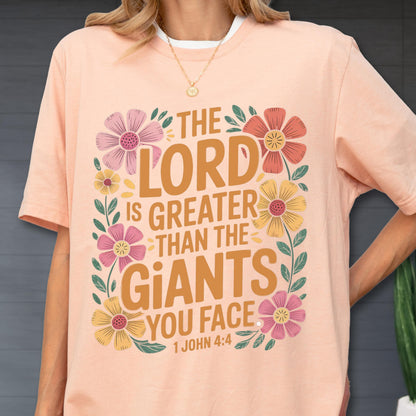 May include: A peach-colored t-shirt featuring the text THE LORD IS GREATER THAN THE GIANTS YOU FACE 1 JOHN 4:4 in brown lettering, surrounded by a decorative border of pink, yellow, and red flowers with green leaves and stems.