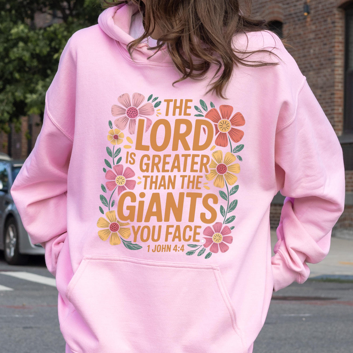 May include: A light pink hoodie with a front pocket and a hood. The back of the hoodie features the text "THE LORD IS GREATER THAN THE GIANTS YOU FACE" in orange, with floral accents. The text is from 1 John 4:4.