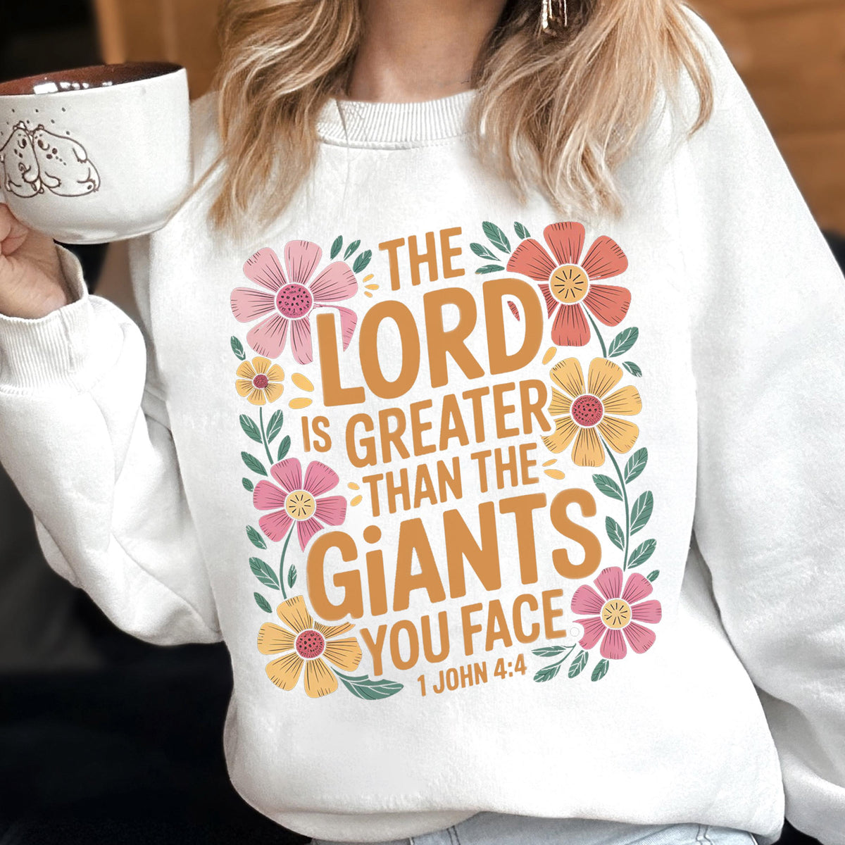 May include: White sweatshirt with the text "THE LORD IS GREATER THAN THE GIANTS YOU FACE" in orange and surrounded by pink, yellow, and orange flowers. The sweatshirt also has the Bible verse 1 John 4:4.