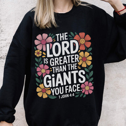 May include: Black sweatshirt with the text "THE LORD IS GREATER THAN THE GIANTS YOU FACE" in white, surrounded by colorful floral designs. The text "1 JOHN 4:4" is at the bottom.
