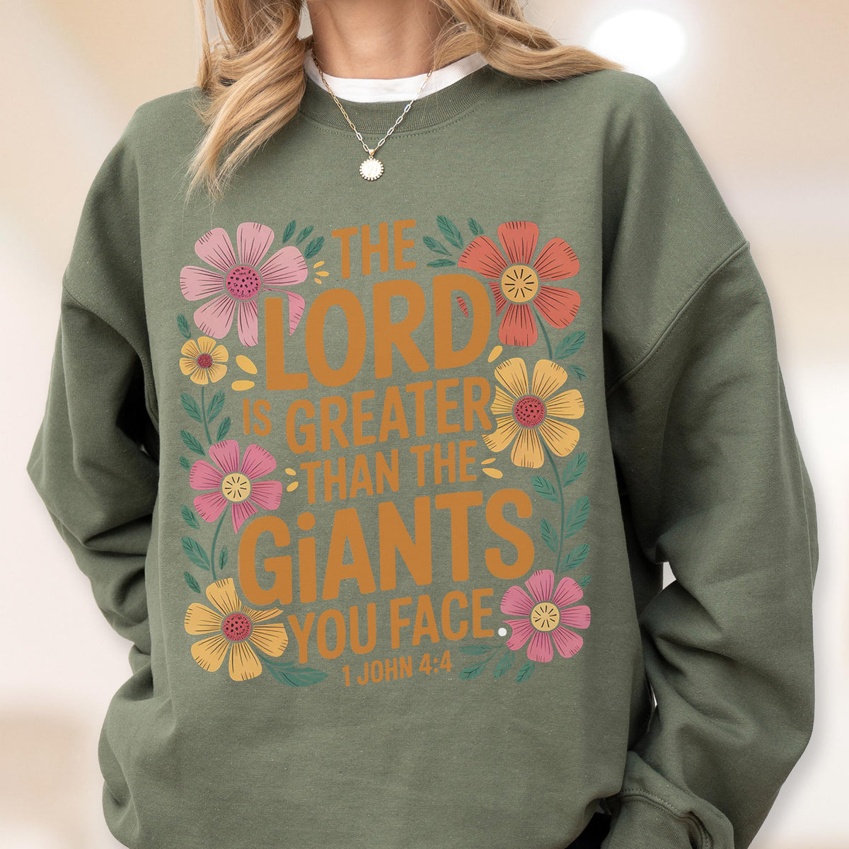May include: Olive green sweatshirt with a floral design and the text "THE LORD IS GREATER THAN THE GIANTS YOU FACE." The design includes pink, orange, and yellow flowers. The text "1 JOHN 4:4" is at the bottom.