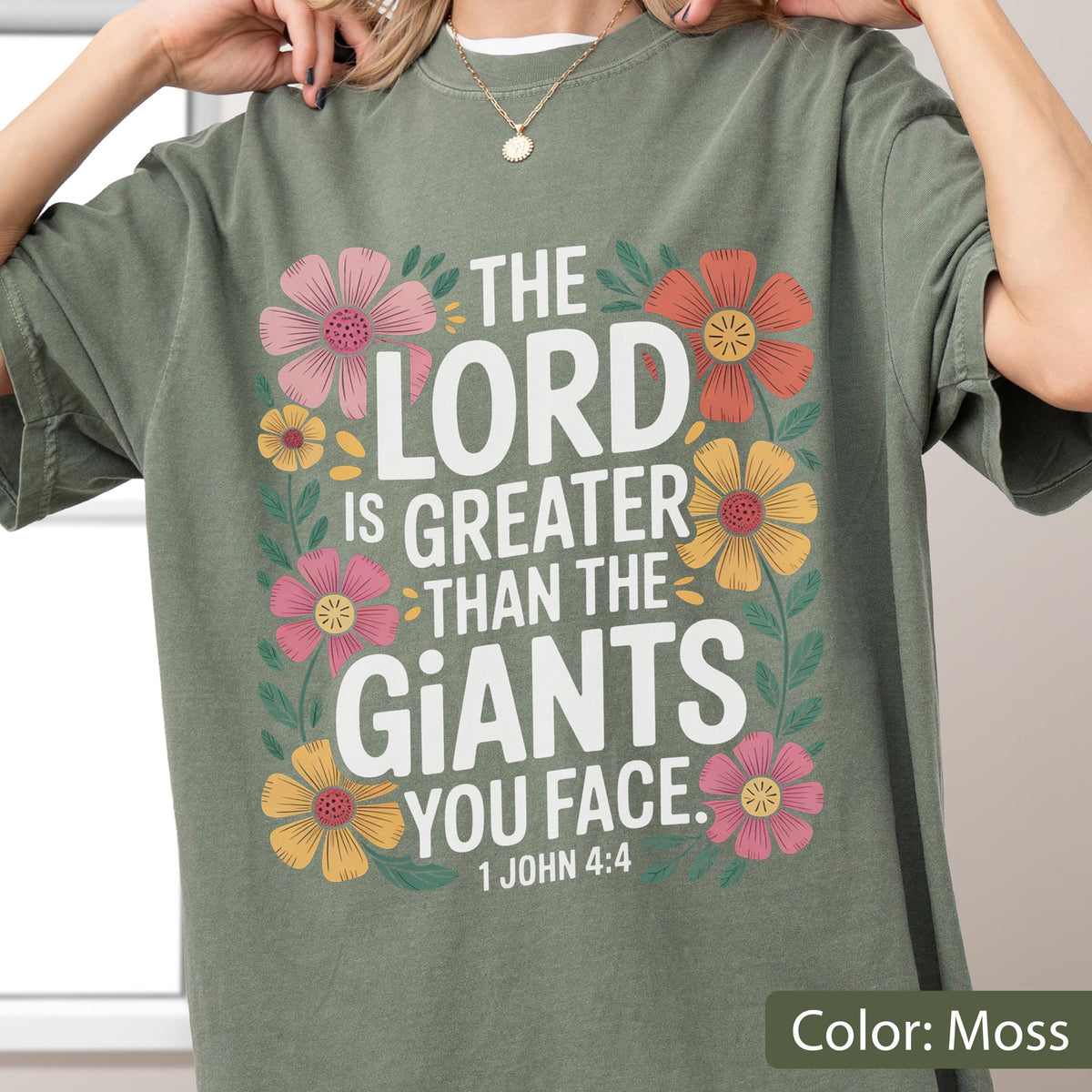 May include: Moss green t-shirt with a floral design and the text "THE LORD IS GREATER THAN THE GIANTS YOU FACE. 1 JOHN 4:4". The design includes pink, orange, and yellow flowers.
