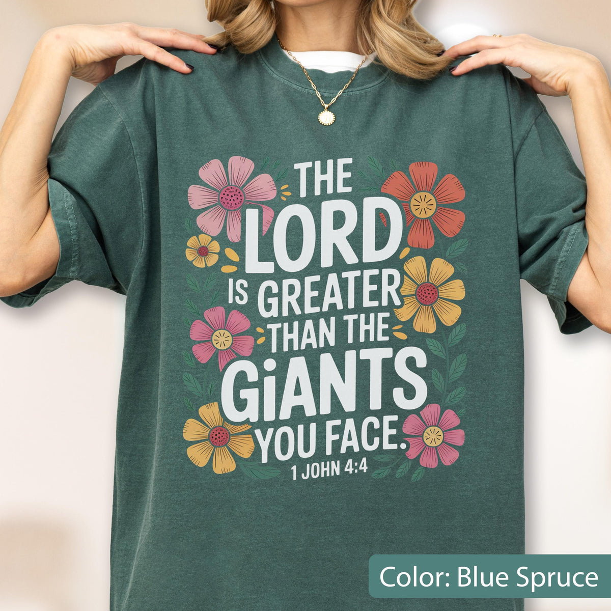 May include: Blue spruce t-shirt with the text "THE LORD IS GREATER THAN THE GIANTS YOU FACE" and the Bible verse 1 John 4:4. The design includes colorful floral accents in pink, orange, and yellow.