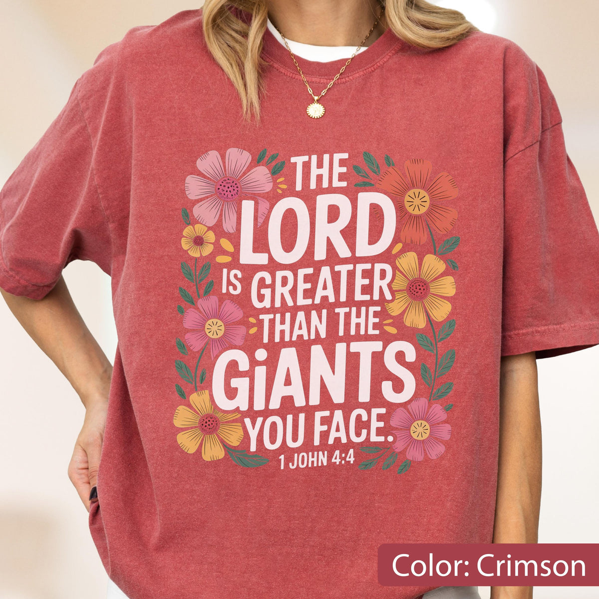 May include: Crimson-colored t-shirt with the text "THE LORD IS GREATER THAN THE GIANTS YOU FACE" surrounded by colorful floral illustrations. The text "1 JOHN 4:4" is below the main text. The shirt is a comfortable, casual style.