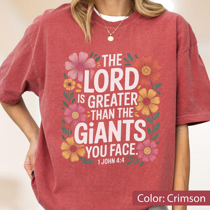 May include: Crimson-colored t-shirt with the text "THE LORD IS GREATER THAN THE GIANTS YOU FACE" surrounded by colorful floral illustrations. The text "1 JOHN 4:4" is below the main text. The shirt is a comfortable, casual style.