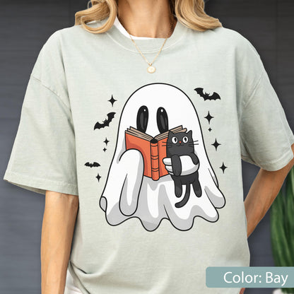 May include: A light green t-shirt featuring a cartoon ghost reading a book and holding a black cat. The design includes bats and stars, with the text "Color: Bay" at the bottom. The shirt is a relaxed fit.