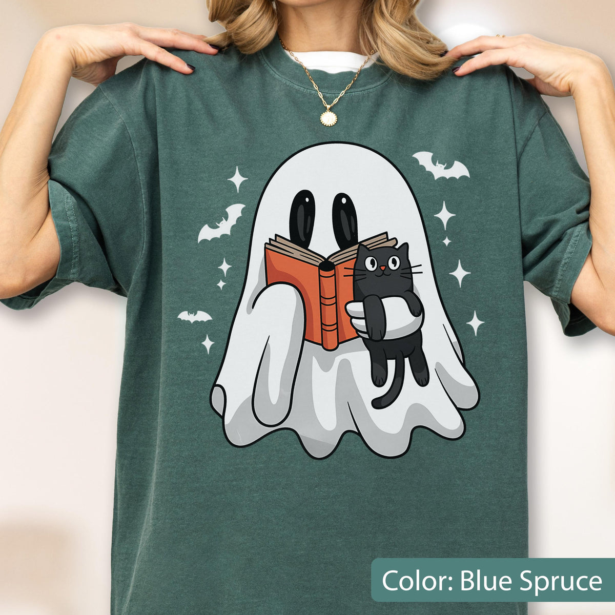 May include: Blue spruce t-shirt featuring a cartoon ghost reading a book with a black cat. The ghost is white with black eyes, holding an orange book. Bats and stars are scattered around the ghost. The text "Color: Blue Spruce" is at the bottom.