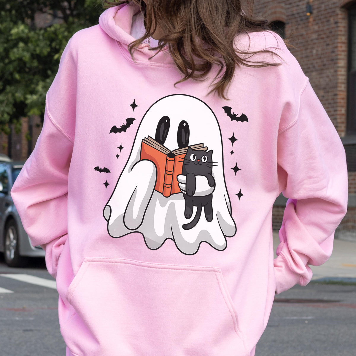 May include: Pink hooded sweatshirt featuring a white ghost reading a book with a black cat. The ghost, cat, bats, and stars are black. The book is orange. The sweatshirt has a front pocket and a hood.