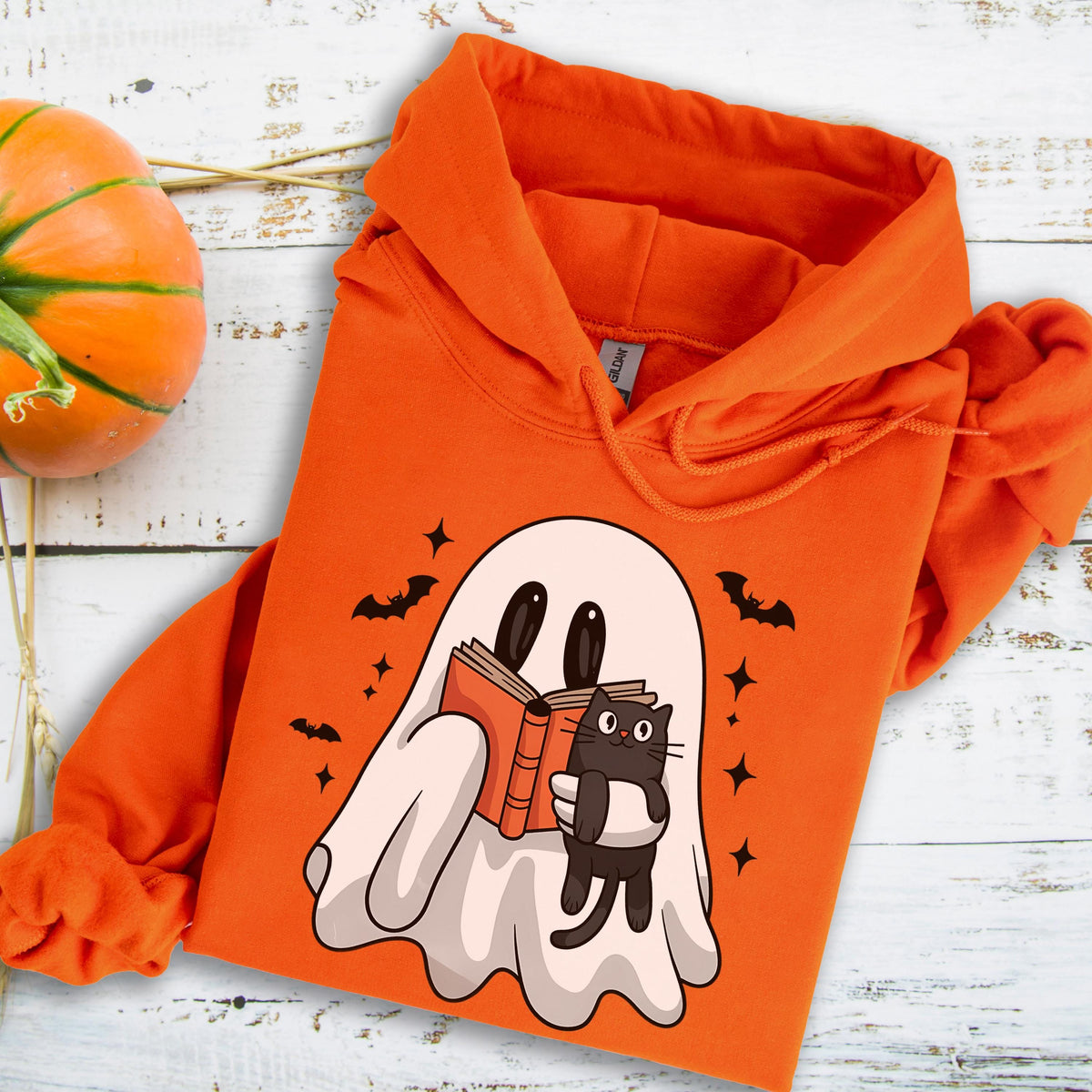 May include: Orange Halloween-themed hoodie featuring a cartoon ghost reading a book and holding a black cat. The design includes bats and stars. A pumpkin is visible in the background. The hoodie is made of a soft material.