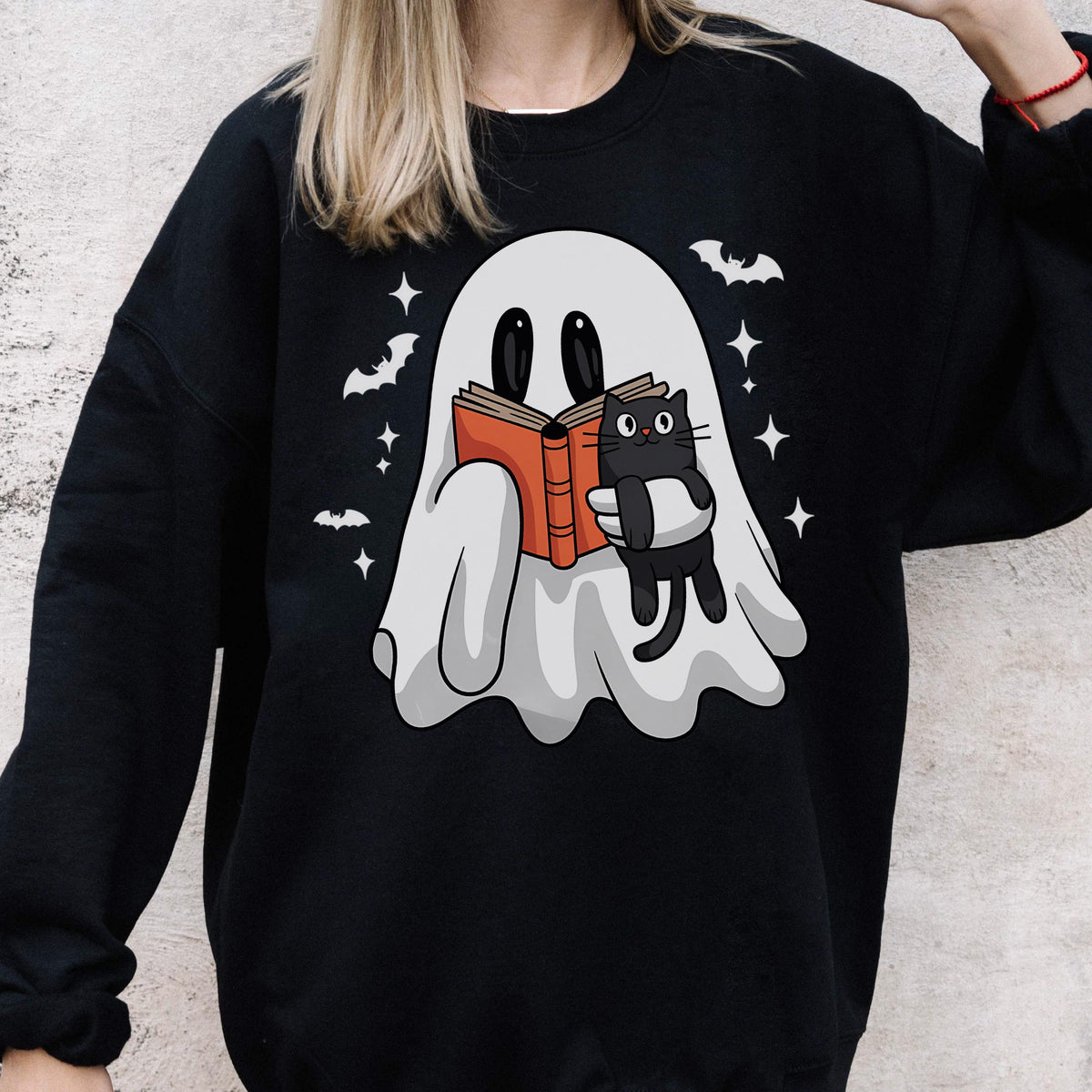 May include: Black sweatshirt featuring a white ghost reading a book with a black cat. The ghost is holding an orange book with the cat in its arms. White bats and stars are scattered around the ghost.