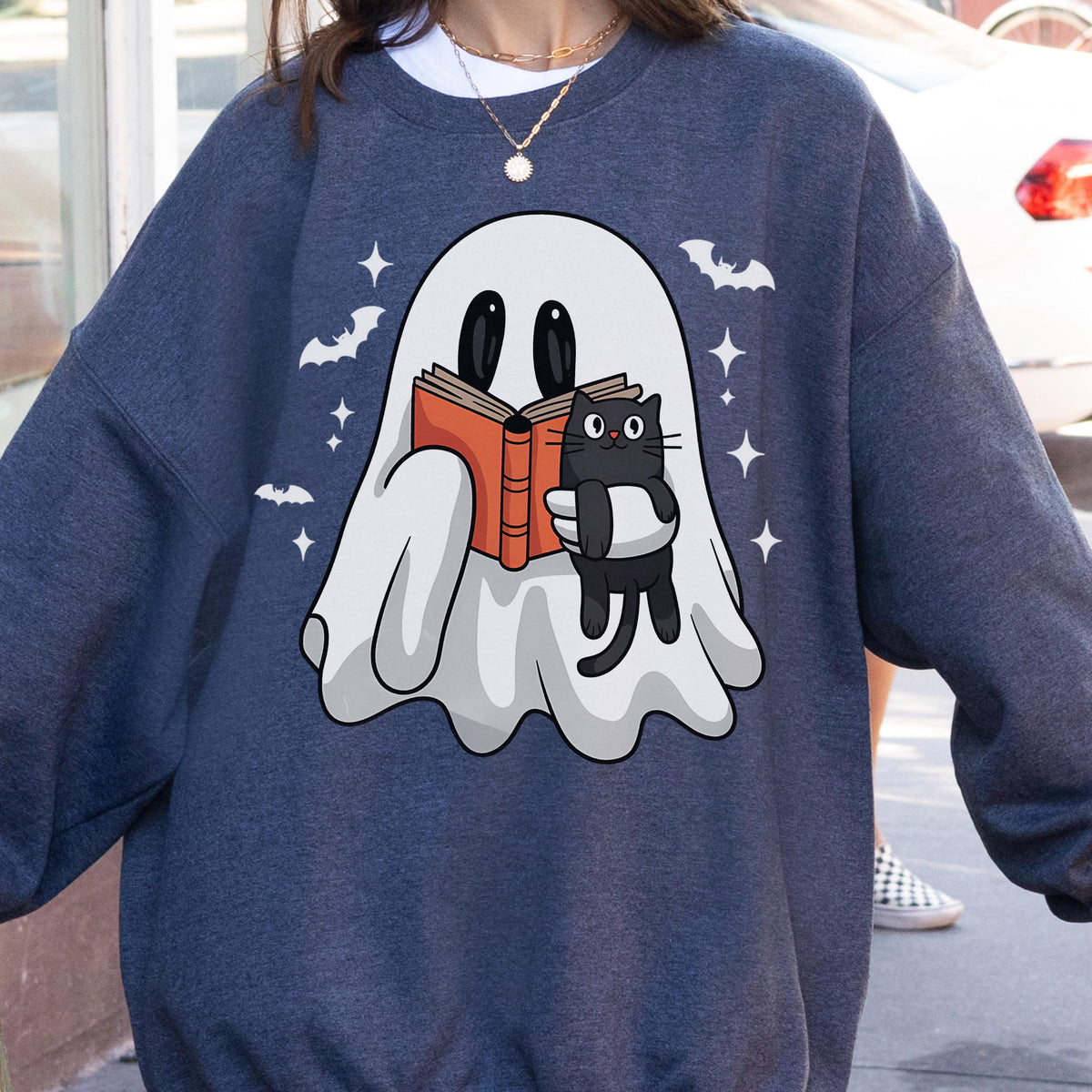 May include: A navy blue sweatshirt featuring a cartoon ghost reading a book with a black cat. The ghost is holding an orange book, and the cat is hugging the book. White bats and stars are scattered around the ghost.