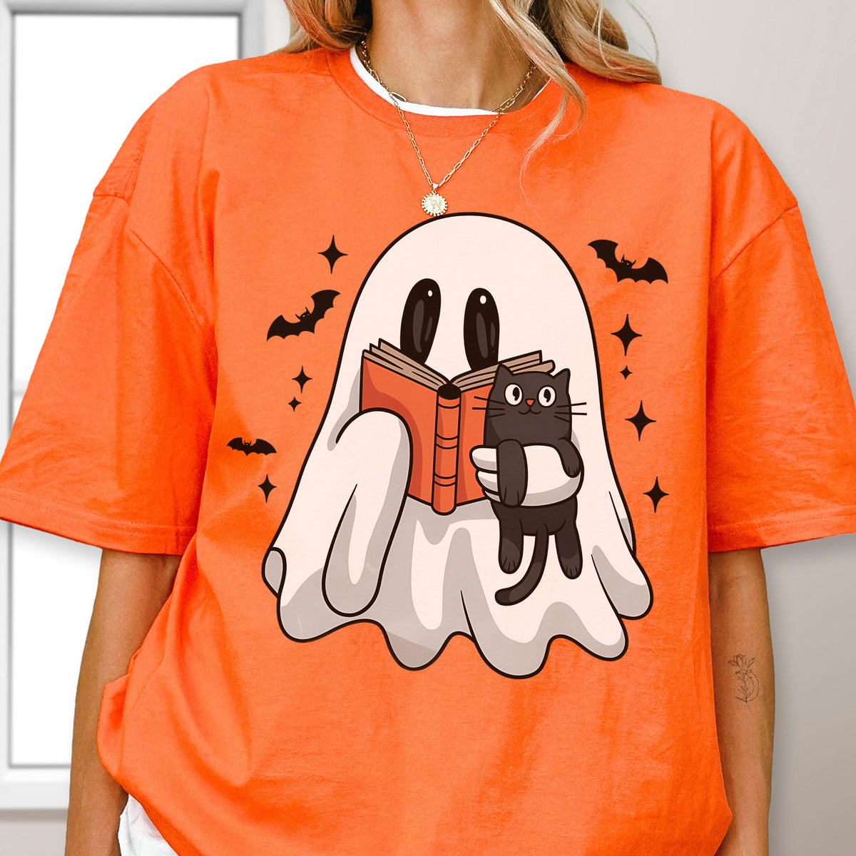 May include: An orange short-sleeve t-shirt featuring a graphic of a white ghost reading an orange book while holding a black cat. The design includes small black bats and four-pointed stars surrounding the ghost, printed on the front of the shirt.