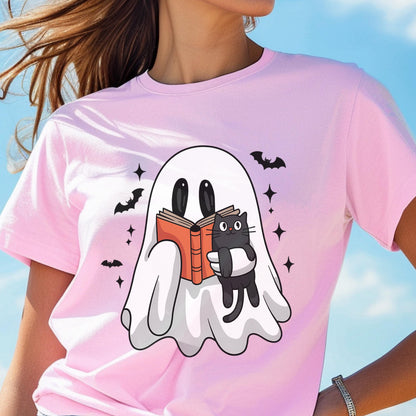 May include: A light pink t-shirt featuring a graphic print of a white ghost holding an open orange book and a small black cat. The design includes several black bats and four-pointed star shapes surrounding the ghost on the front of the shirt.