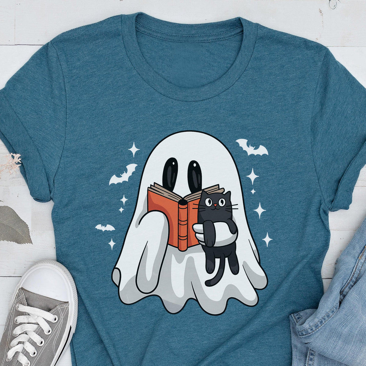 May include: A teal crew neck t-shirt featuring a graphic of a white ghost holding an open orange book and a small black cat. The design includes white stars and silhouettes of flying bats surrounding the ghost on the front of the garment.