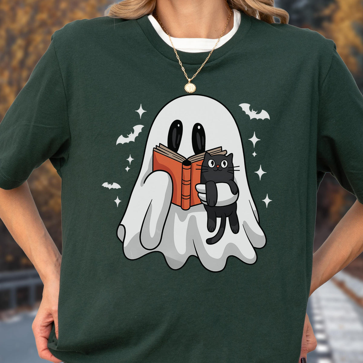 May include: A dark green t-shirt featuring a graphic of a white ghost holding an open orange book and a small black cat. The design includes small white stars and bat silhouettes surrounding the ghost, printed on the front of the garment.