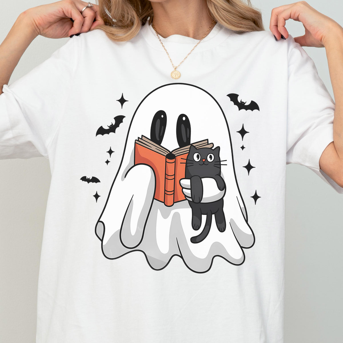 May include: A white t-shirt featuring a graphic of a white ghost reading an orange book while holding a black cat. The design includes small black bats and four-pointed star shapes surrounding the ghost on the front of the garment.