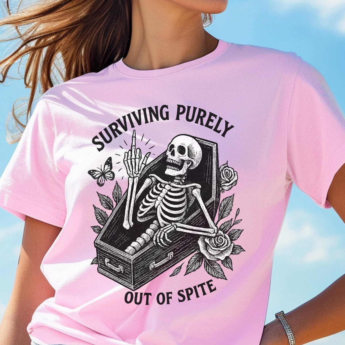 May include: A light pink t-shirt featuring a black graphic of a skeleton inside a coffin, giving the middle finger. The design includes roses and a butterfly, with the text "SURVIVING PURELY OUT OF SPITE" arched around the central illustration.