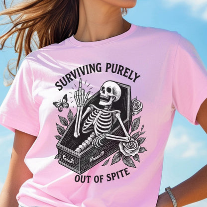 May include: A light pink t-shirt featuring a black graphic of a skeleton inside a coffin, giving the middle finger. The design includes roses and a butterfly, with the text "SURVIVING PURELY OUT OF SPITE" arched around the central illustration.