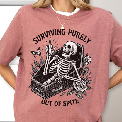 May include: A dusty rose t-shirt featuring a black graphic of a skeleton inside a coffin, giving the middle finger. The text "SURVIVING PURELY" is arched above the coffin, and "OUT OF SPITE" is printed below, surrounded by roses and a butterfly.